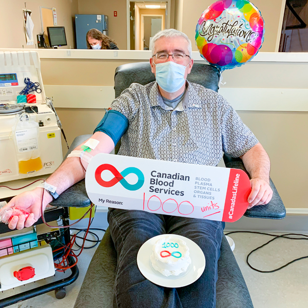 Image of blood donor Allen Veal holding up a My Reasons sign while sitting in a chair donating blood at the Charlottetown, PEI donor centre