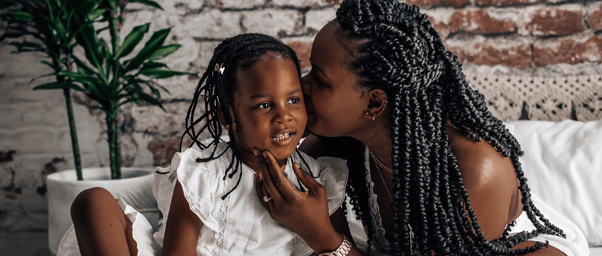 A mother gives her young daughter a kiss on the cheek, sitting on a bed in front of a brick wall. 